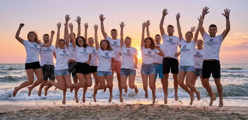 Grup de tineri entuziasmați sărind pe plaja din Costinești la apus, purtând tricouri albe personalizate cu logo-ul Casa Duca.